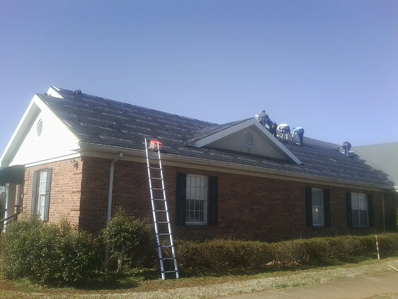Crew tearing shingles off a brick ranch roof with a ladder set — Emergency Roof Repair work by our crew in Glen Allen